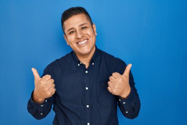 Latin kökenli genç adam mavi arka plan başarı işaretinin üzerinde duruyor. Elleriyle olumlu jest yapıyor. Gülümsüyor ve mutlu. Neşeli ifade ve kazanan jest. 
