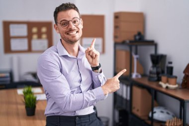 Ofisteki genç İspanyol adam gülümsüyor ve kameraya bakıyor. İki eliyle ve parmaklarıyla kenarı işaret ediyor.. 