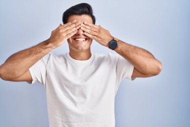 Mavi arka planda dikilen İspanyol adam elleri gülücüklü ve komik bir şekilde gözlerini kapatıyor. kör kavram. 