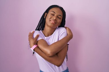 Örgü örgülü Afro-Amerikan kadın pembe arka planda durup kendine sarılıyor mutlu ve pozitif, kendinden emin gülümsüyor. Sevgi ve özgüven. 