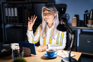 Gri saçlı orta yaşlı bir kadın gece ofiste çalışıyor ve dört numaralı parmaklarıyla işaret ediyor. Kendinden emin ve mutlu bir şekilde gülümsüyor.. 