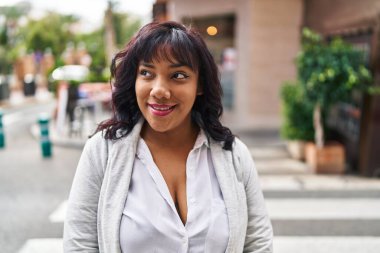 Genç, güzel, Latin bir kadın gülümsüyor. Kendinden emin, caddeye bakıyor.