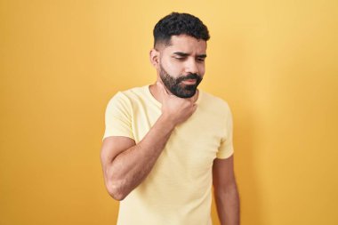Sarı arka planda sakallı İspanyol bir adam ağrılı boynuna dokunuyor, grip, mantar ve enfeksiyon yüzünden boğazı ağrıyor. 