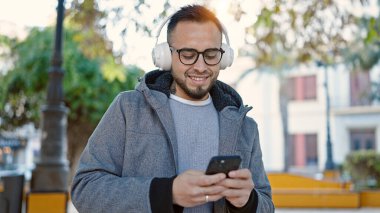 İspanyol adam, parkta akıllı telefon kullanarak müzik dinlerken kendinden emin gülümsüyor.