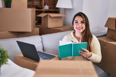 Genç ve güzel İspanyol kadın dizüstü bilgisayar kullanıyor. Yeni evinde deftere yazıyor.