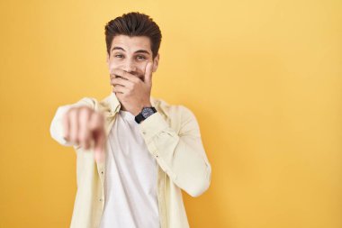 Sarı arka planda dikilen İspanyol genç adam sana gülüyor, eli ağzında kamerayı işaret ediyor, utanç ifadesi. 