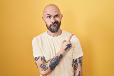 Sarı arka planda dikilen İspanyol dövmeli adam endişeli, endişeli ve şaşırmış ifadesini işaret ediyor. 