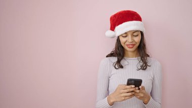 Genç, güzel, İspanyol bir kadın noel şapkası takıyor ve pembe arka plan üzerinde akıllı telefon kullanıyor.