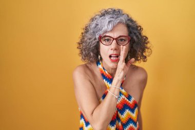 Orta yaşlı bir kadın, sarı arka planda dikilmiş, ağzı açık, gizli söylentiler yayıyor, kötü niyetli konuşmalar fısıldıyor. 