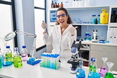 Young hispanic woman working at scientist laboratory pointing thumb up to the side smiling happy with open mouth 