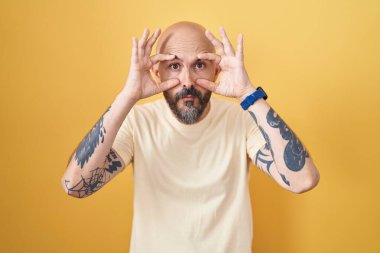 Sarı arka planda dikilen İspanyol dövmeli adam parmaklarıyla gözlerini açmaya çalışıyor, uykulu ve yorgun bir şekilde sabah yorgunluğu çekiyor. 