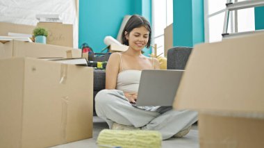 Young beautiful hispanic woman using laptop sitting on floor at new home