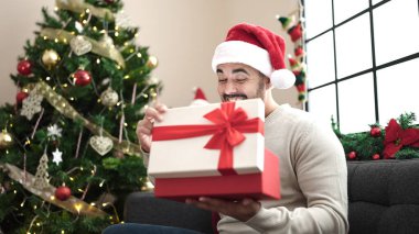 Genç İspanyol adam evdeki Noel ağacının yanında kanepeye oturmuş hediyesini açıyor.