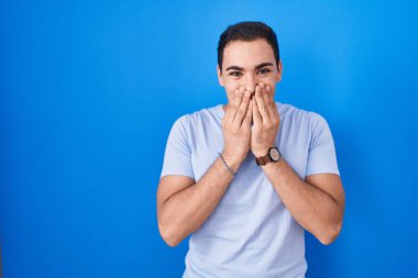 Mavi arka planda dikilen Latin asıllı genç adam gülüyor ve utanıyor kıkırdıyor elleriyle ağzını kapatıyor, dedikodu ve skandal konsepti yapıyor. 