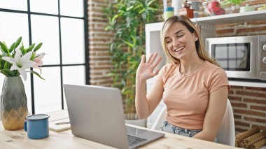 Masada oturan genç sarışın kadın yemek odasında video görüşmesi yapıyor.