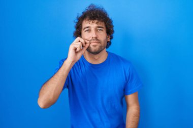 Hispanic young man standing over blue background mouth and lips shut as zip with fingers. secret and silent, taboo talking 