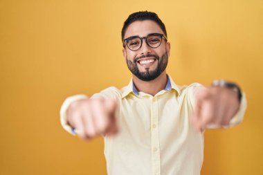 İş kıyafetleri ve gözlük takan İspanyol genç adam seni ve kamerayı işaret ediyor, pozitif ve neşeli gülümsüyor. 