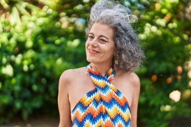 Orta yaşlı, kır saçlı, gülümseyen, kendine güvenen bir kadın parkın kenarına bakıyor.