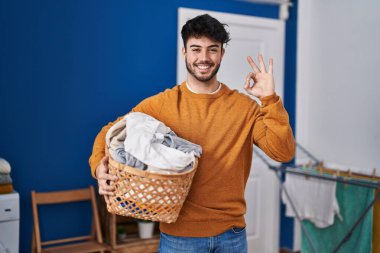 Çamaşır odasında sakallı İspanyol bir adam çamaşır sepetini tutuyor parmaklarıyla imza atıyor, gülümseyerek mükemmel bir sembol oluşturuyor. 