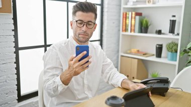 Ciddi yüzlü İspanyol iş adamı, ofiste akıllı telefon kullanıyor, zarif giyinmiş başarılı bir işçinin portresi, kapalı alanda konsantre oluyor.