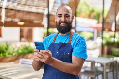 Young bald man waiter smiling confident using smartphone at coffee shop terrace