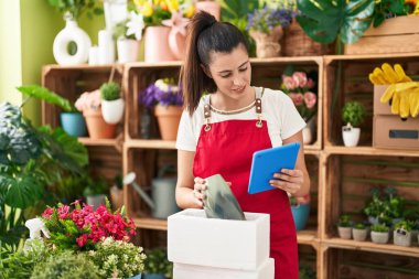 Young beautiful hispanic woman florist unpacking plant jar using touchpad at flower shop