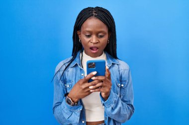 Örgülü Afrikalı bir kadın akıllı telefon kullanarak mesaj yazıyor. Korkmuş ve ağzı açık bir şekilde şaşırmış, inançsız bir yüz. 