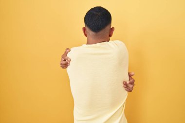 Sarı arka planda dikilen İspanyol genç adam kendine sarılıyor mutlu ve pozitif bir şekilde. Sevgi ve özgüven. 