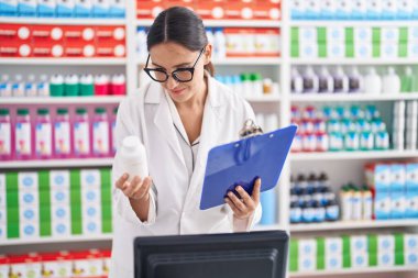 Genç ve güzel İspanyol kadın eczacı elinde ilaç şişesiyle eczanede doküman okuyor.