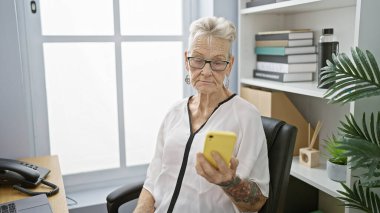 Zarif, gri saçlı kıdemli kadın patron, işine konsantre oluyor, ofis karmaşasının ortasında akıllı telefondan ciddi mesajlar yazıyor. Yaş ve teknolojinin mükemmel bir karışımı..