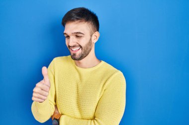 Hispanic man standing over blue background looking proud, smiling doing thumbs up gesture to the side 