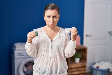 Orta yaşlı İspanyol kadın çamaşır deterjanı ve çamaşır deterjanı tutuyor şüpheci ve gergin, sorun yüzünden surat asıyor. negatif kişi. 