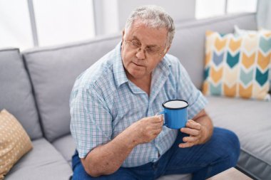Orta yaşlı, kır saçlı, kahve içen adam evdeki kanepede oturuyor.