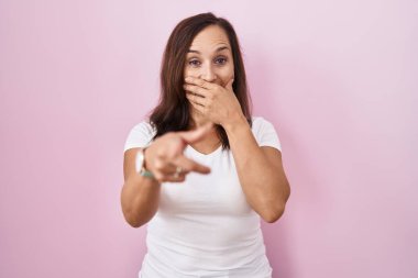 Orta yaşlı esmer kadın pembe arka planda durup sana gülüyor, eli ağzında kamerayı işaret ediyor, utanç ifadesi var. 