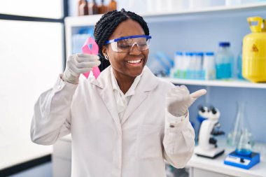 Laboratuvarda çalışan Afrikalı Amerikalı bir kadın elinde pembe kurdeleyle ağzı açık gülümsüyor. 