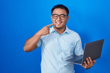 Çinli genç adam bilgisayarda gülümseyip parmaklarını ve ağzını göstererek gülümsüyor. diş sağlığı kavramı. 