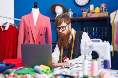 Young redhead man tailor using laptop at clothing factory