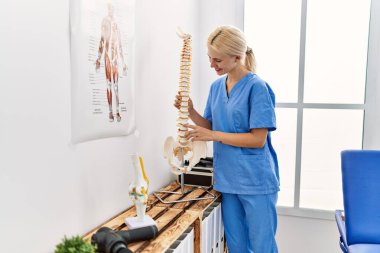 Genç sarışın kadın pisiyoterapist, rehabilitasyon kliniğinde kendine güvenen, omurgaya dokunan, kendinden emin bir şekilde gülümsüyor.