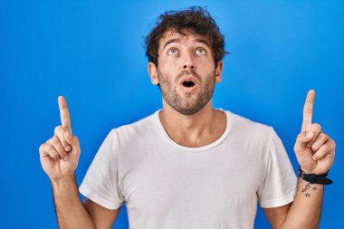 Mavi arka planda duran İspanyol genç adam şaşırmış ve şaşırmıştı. Yukarı baktı ve parmaklarıyla işaret etti ve kollarını kaldırdı.. 