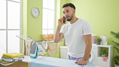 Young arab man talking on smartphone ironing clothes at laundry room