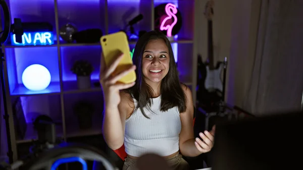 Smiling young hispanic woman, streaming a thrilling video game, engrossed in digital entertainment from her neon-lit gaming room during a lively video call