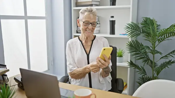 オフィスでスマートフォンに陽気なメッセージを入力するシニアグレー髪のビジネス女性の笑顔 — ストック写真