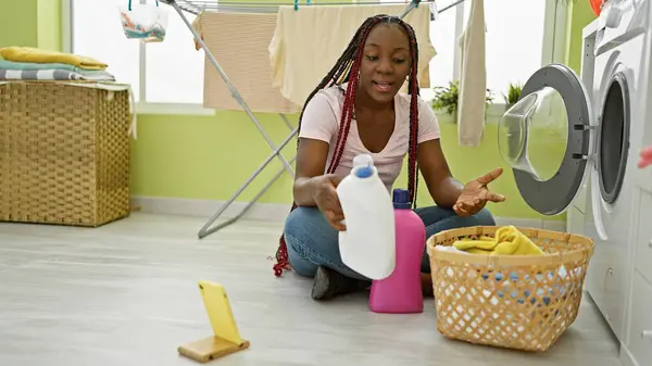 Charming african american woman, her braids tied up, beautifully chatting on a video call, gleefully showcasing her laundry detergent right from her neat laundry room while doing house chores