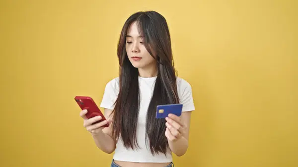 Young chinese woman shopping with smartphone and credit card with relaxed expression over isolated yellow background