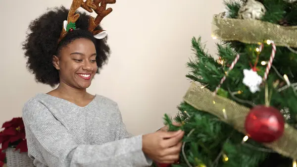 African american woman wearing reindeer ears decorating christmas tree at home