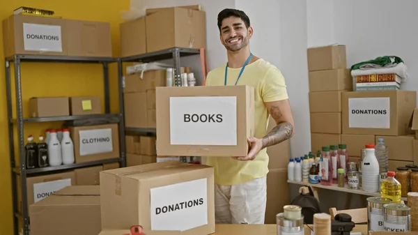 Young hispanic man volunteer holding books package to donate smiling at charity center