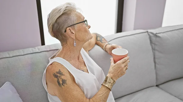 Comfortable grey-haired senior woman relaxing with a cup of coffee on her sofa at home