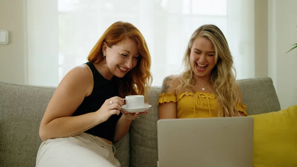 Two women drinking coffee using laptop at home