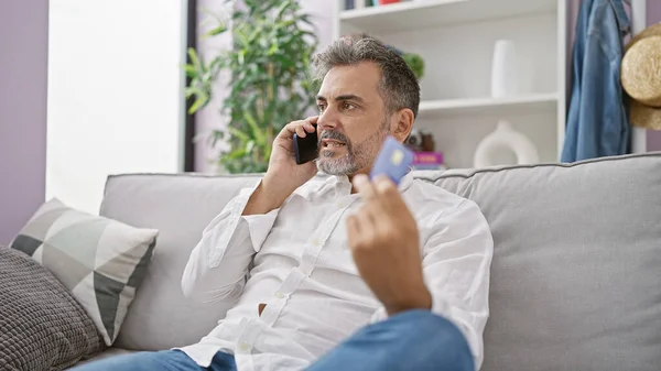 Upset hispanic man with grey hair in a problematic phone talk, holding a credit card at home, expressing worry indoors.