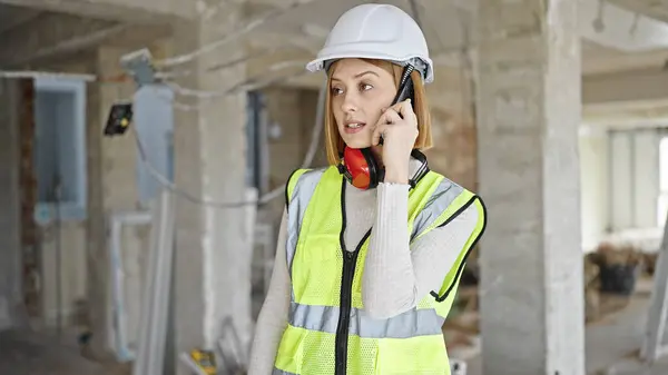 Young blonde woman architect talking on smartphone at construction site